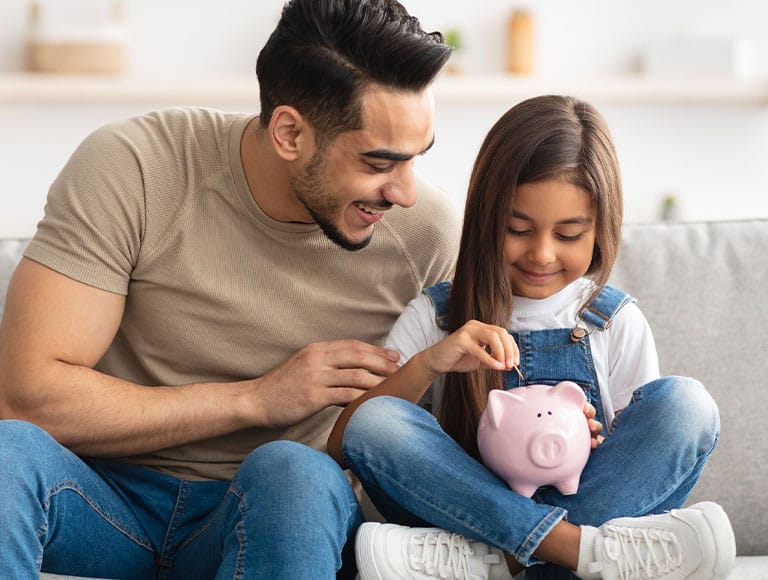 Father and daughter smiling, saving money in a pink piggy bank on a couch.