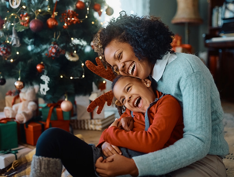 A woman and child laughing together in front of a Christmas tree with presents.