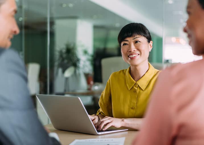 A woman in yellow smiling during a meeting with two colleagues in an office setting.