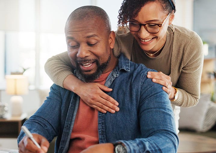 A woman embraces a smiling man seated at a table with papers.