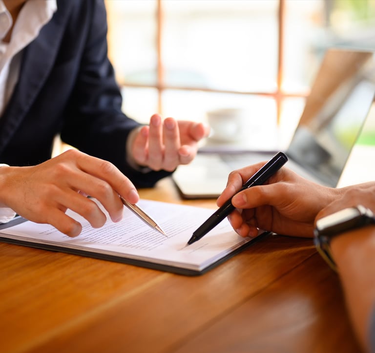 Two people discussing a document with pens, seated at a wooden table with a laptop visible in the background.