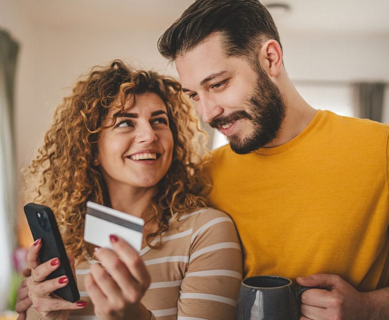 A couple smiling while shopping online with a phone and credit card.
