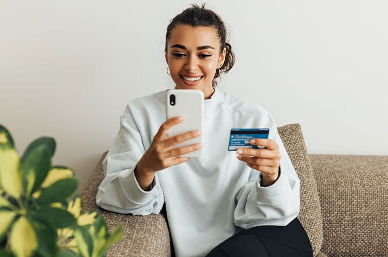 A woman smiles at her smartphone while holding a credit card, sitting on a sofa.