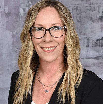 A woman with long blonde hair and glasses smiles in front of a gray background.